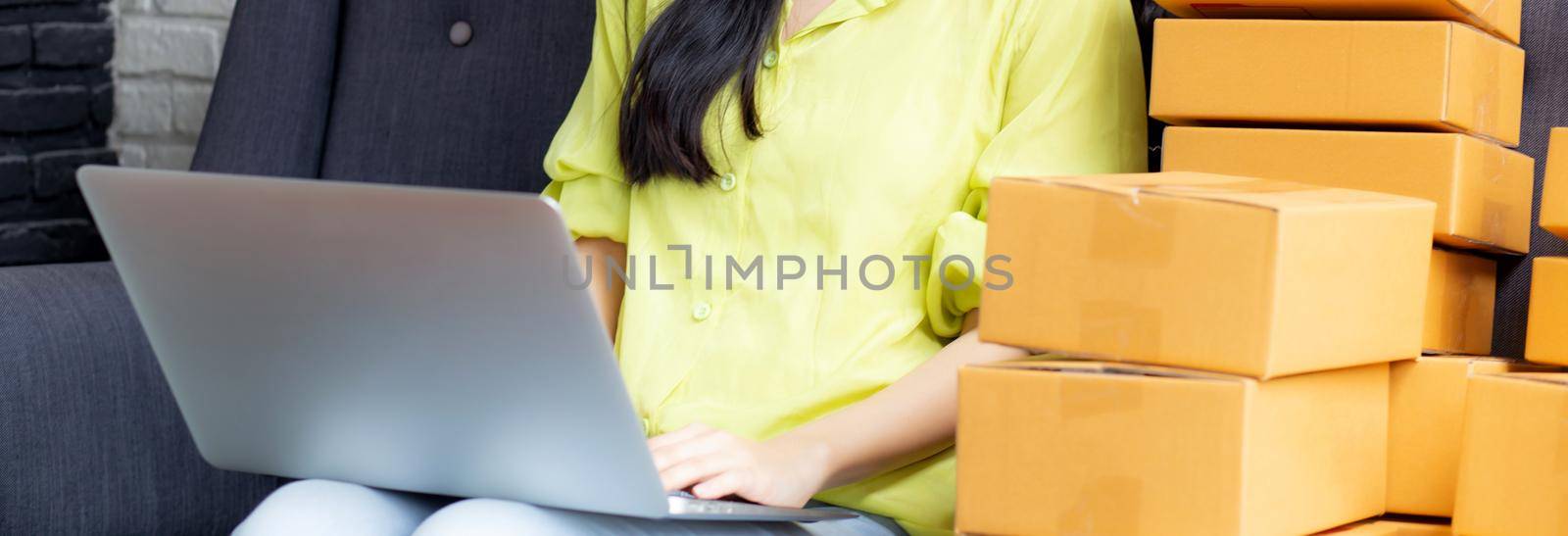 Young asian woman is merchant using laptop computer on sofa checking order of customer with purchase online shopping, business SME or startup, entrepreneur or seller and service delivery product.