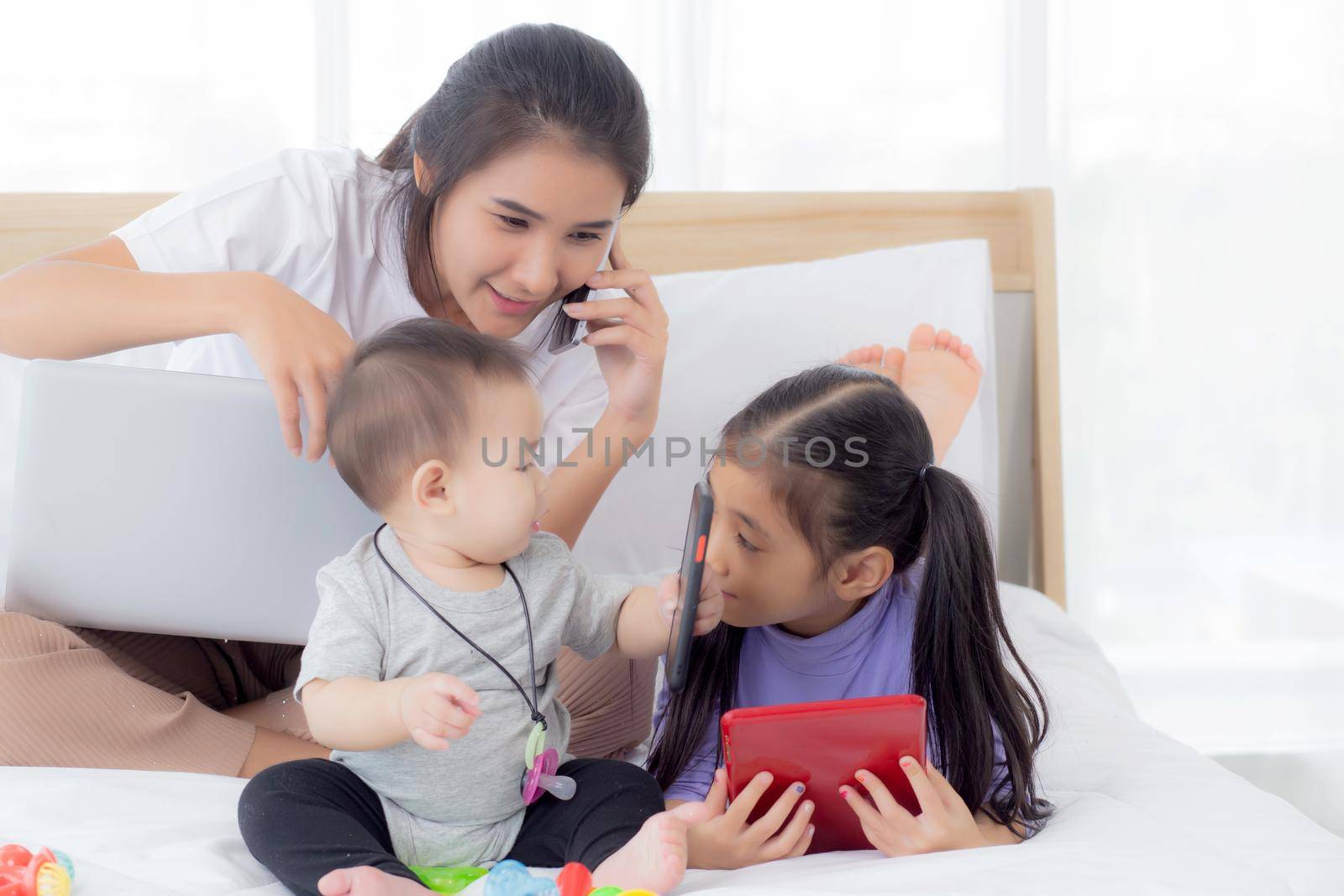 Young asian mother working at home online to internet with laptop computer and talking phone and daughter looking tablet on bed at bedroom, business woman is freelance, new normal, family concept.