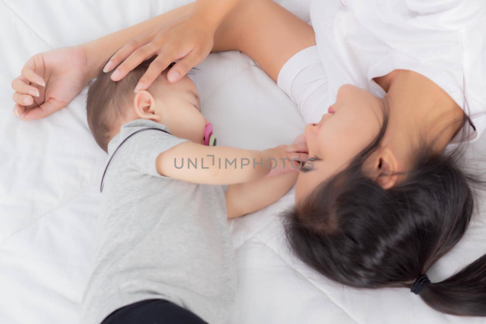Young asian mother touch head of little baby girl with tender on bed in the bedroom, mom love newborn and care, woman with expression with child together, parent and daughter, family concept.