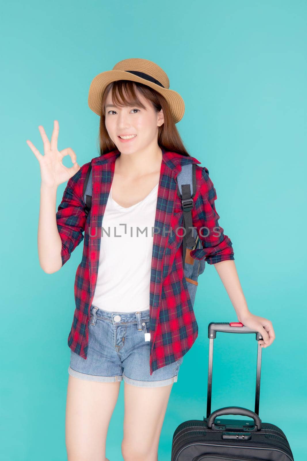 Beautiful young asian woman pulling suitcase isolated on blue background, asia girl cheerful holding luggage walking and gesture ok in vacation with excited, journey and travel concept.