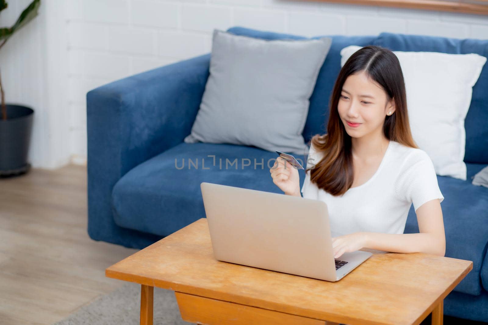 Young asian woman sit on sofa using laptop computer shopping online with credit card buying to internet, happy girl payment with e-business on couch, purchase and payment, business concept.
