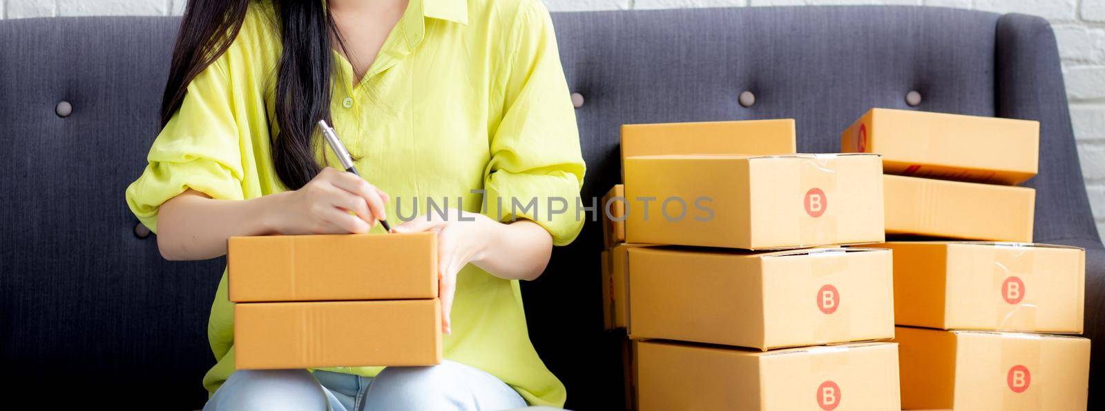 Young asian woman writing address on parcel box at home for delivery order to customer, shipping and logistic, merchant online and seller, business owner or SME, online shopping and e-commerce.