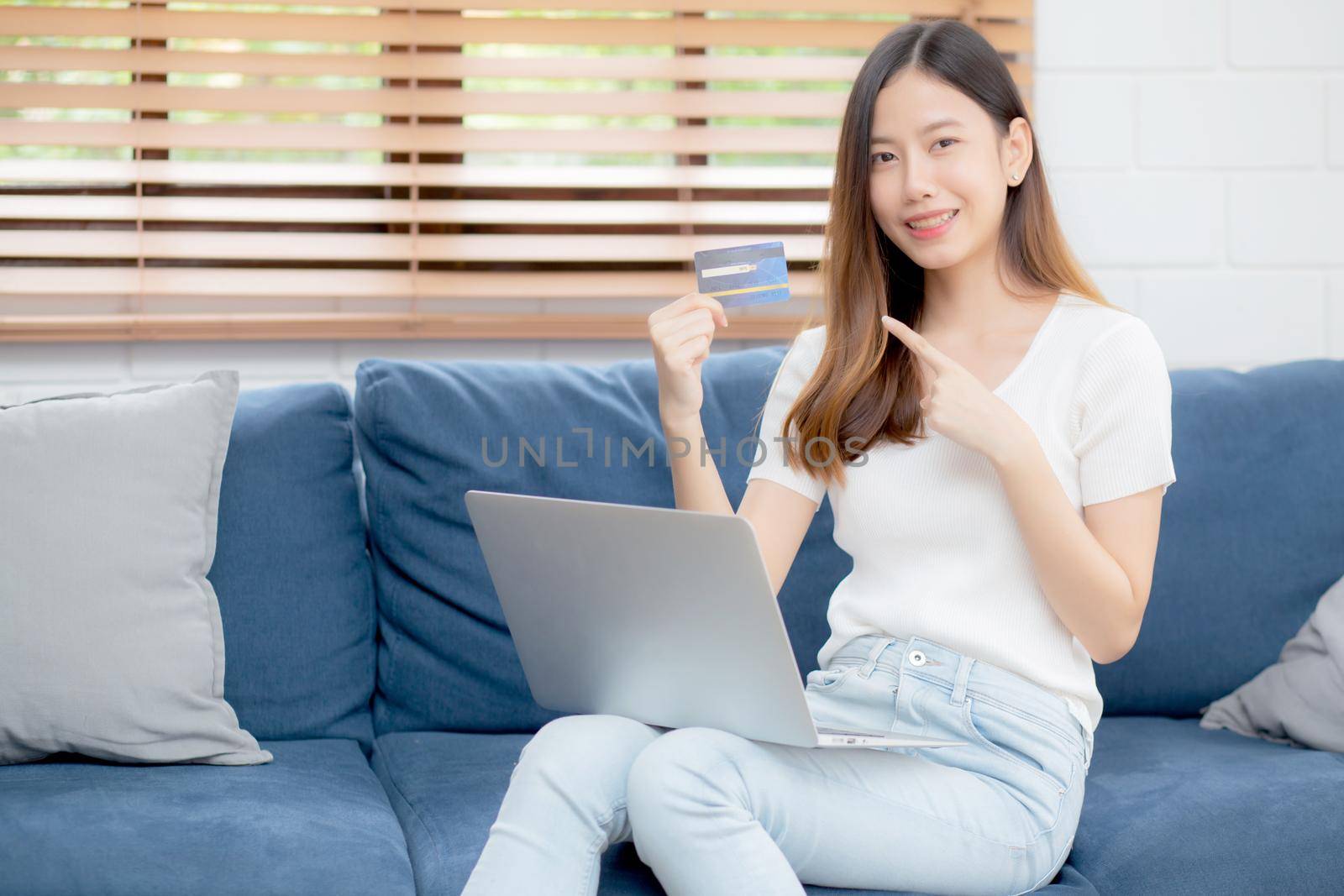Young asian woman sit on sofa using laptop computer shopping online with credit card buying to internet, happy girl payment with e-business on couch, purchase and payment, business concept.