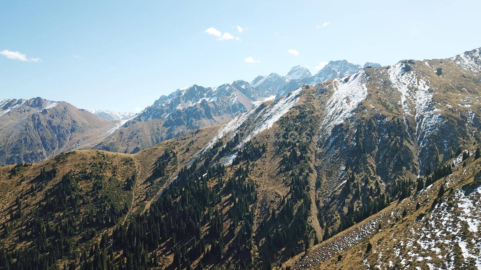 Autumn mountains covered with snow in places. View from the drone, from above. Huge slopes of the gorge, yellow grass and snow. Large stones visible in the river. Shadow of clouds on the ground.