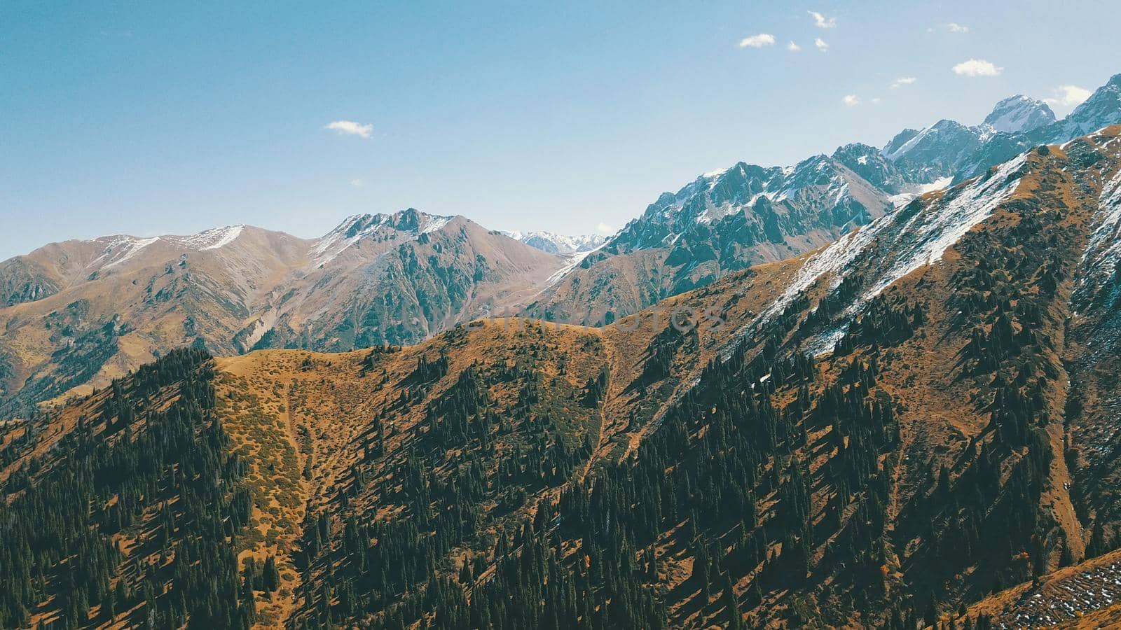 Autumn mountains covered with snow in places. View from the drone, from above. Huge slopes of the gorge, yellow grass and snow. Large stones visible in the river. Shadow of clouds on the ground.