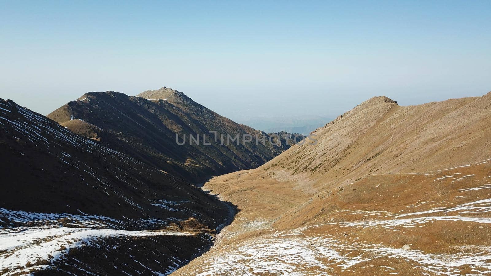 Autumn mountains covered with snow in places. View from the drone, from above. Huge slopes of the gorge, yellow grass and snow. Large stones visible in the river. Shadow of clouds on the ground.