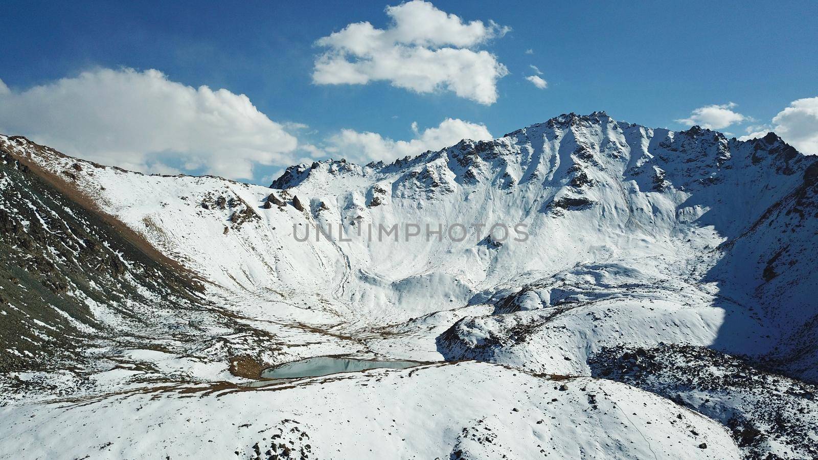 Snow-capped mountains and mountain lake. Top view from a drone. Blue sky and clouds are reflected in the water. Above the mountains. Steep slopes and rocky peaks. The gorge is covered with snow.