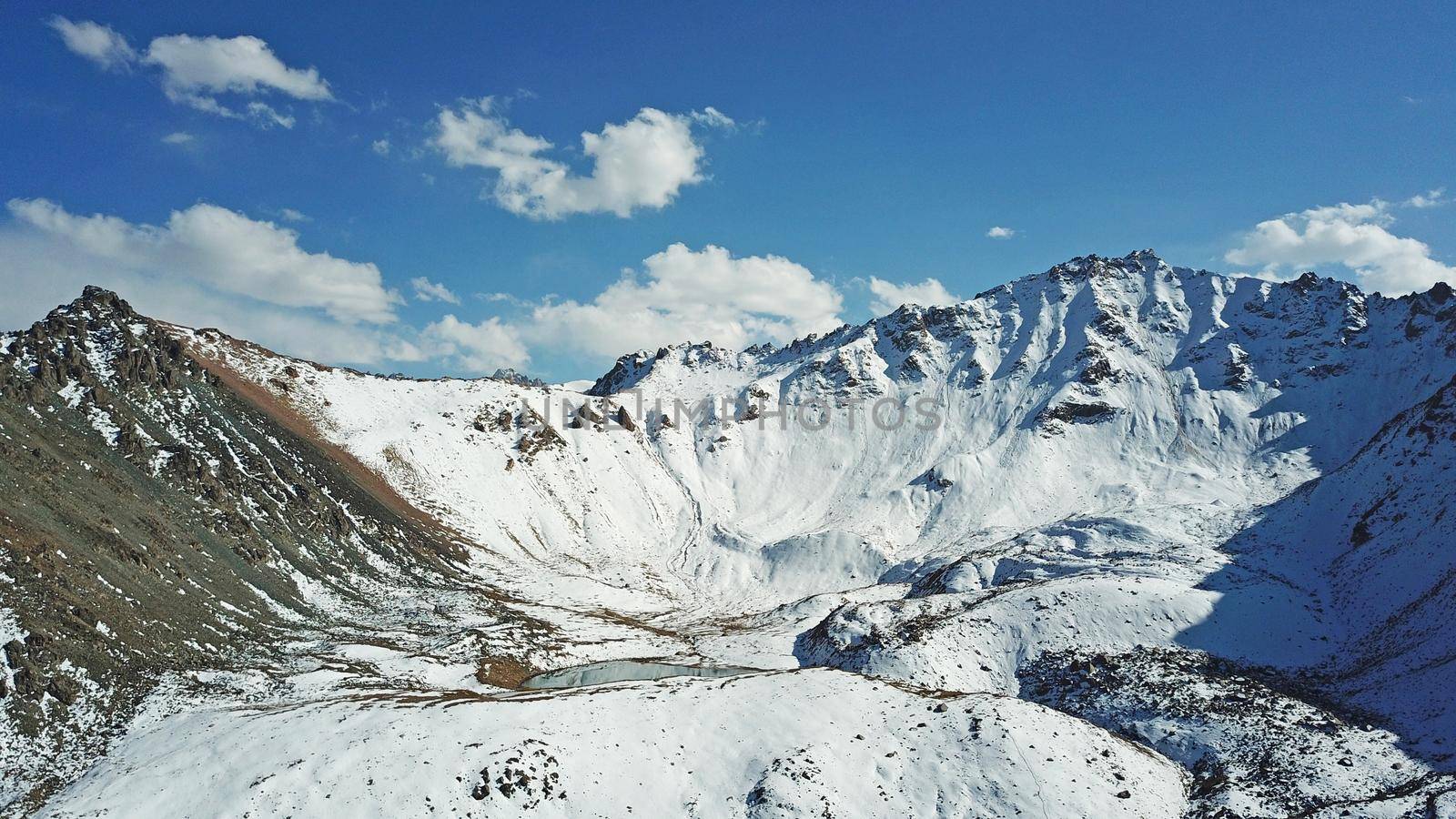 Snow-capped mountains and mountain lake. Top view from a drone. Blue sky and clouds are reflected in the water. Above the mountains. Steep slopes and rocky peaks. The gorge is covered with snow.