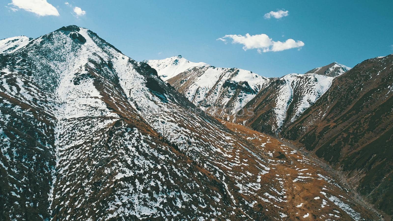 Autumn mountains covered with snow in places. View from the drone, from above. Huge slopes of the gorge, yellow grass and snow. Large stones visible in the river. Shadow of clouds on the ground.