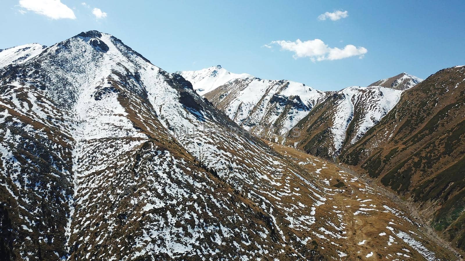 Autumn mountains covered with snow in places. View from the drone, from above. Huge slopes of the gorge, yellow grass and snow. Large stones visible in the river. Shadow of clouds on the ground.