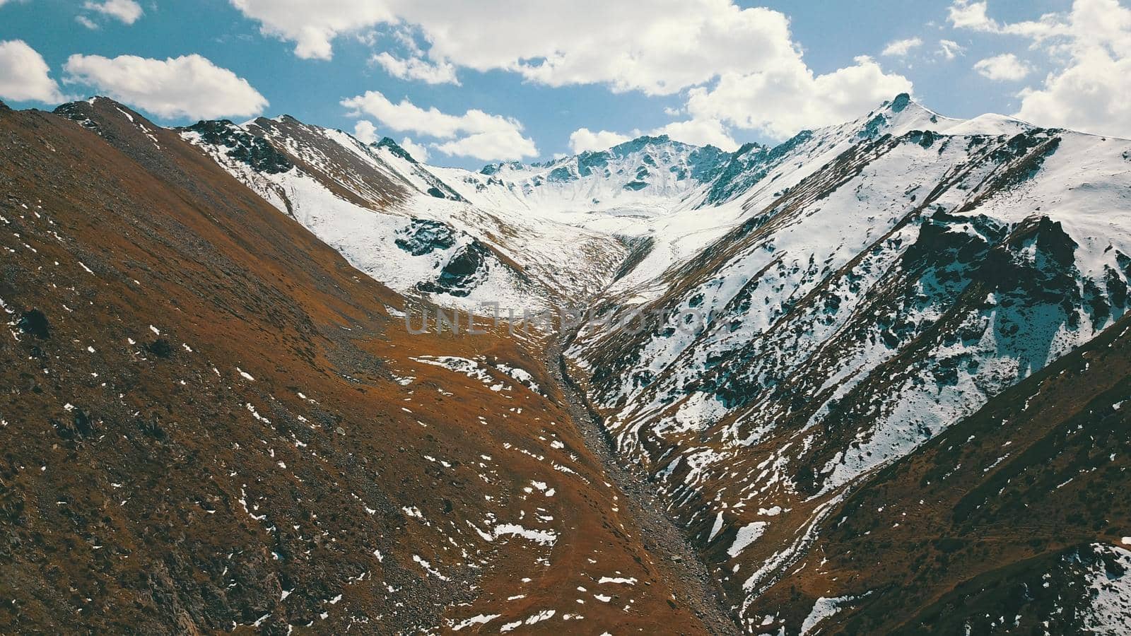 Autumn mountains covered with snow in places. View from the drone, from above. Huge slopes of the gorge, yellow grass and snow. Large stones visible in the river. Shadow of clouds on the ground.