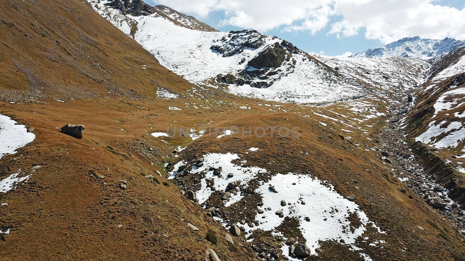 Autumn mountains covered with snow in places. View from the drone, from above. Huge slopes of the gorge, yellow grass and snow. Large stones visible in the river. Shadow of clouds on the ground.