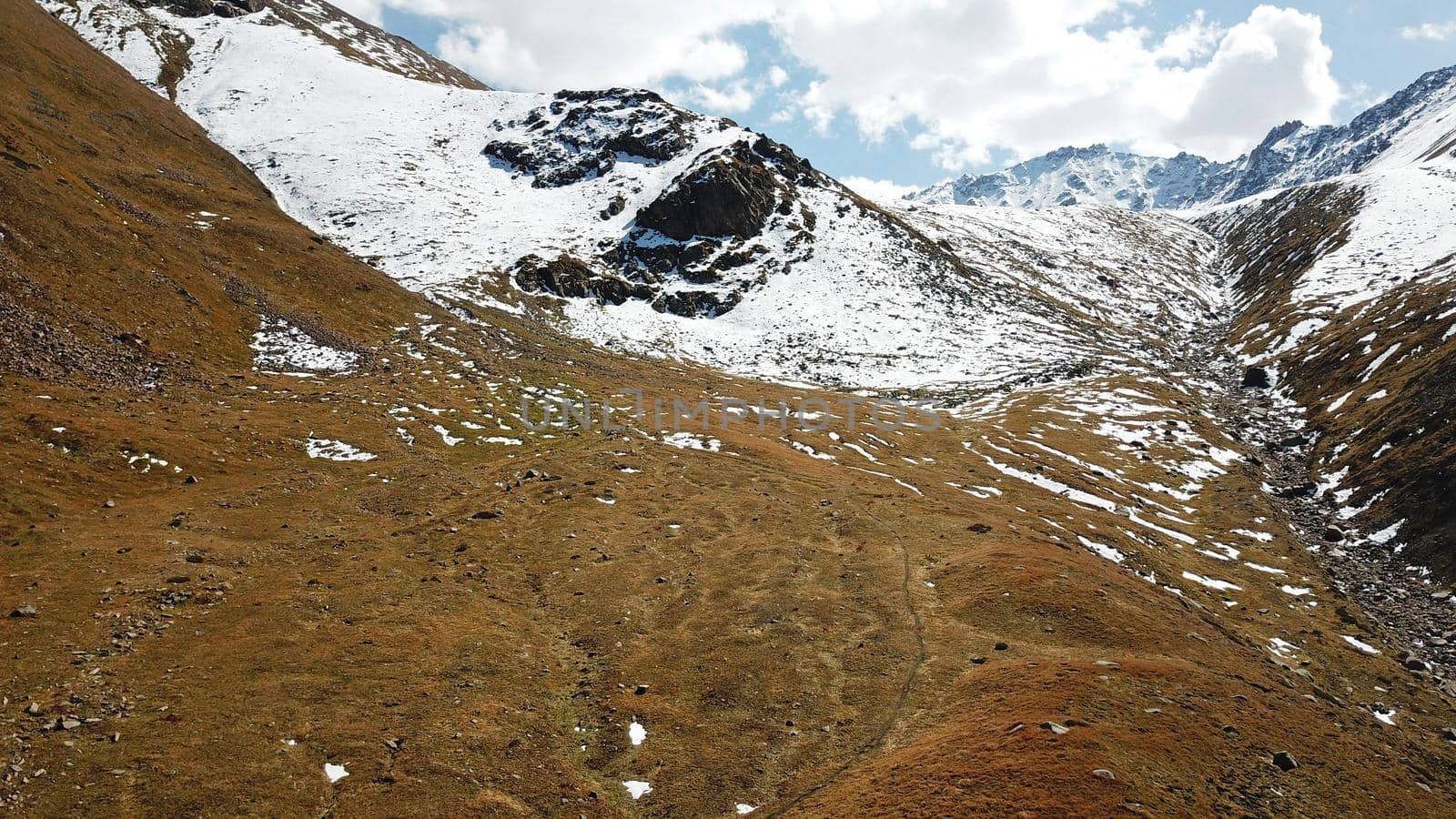 Autumn mountains covered with snow in places. View from the drone, from above. Huge slopes of the gorge, yellow grass and snow. Large stones visible in the river. Shadow of clouds on the ground.