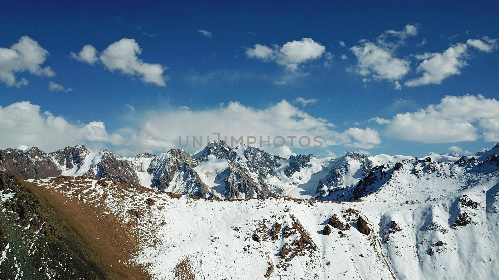 Snow-capped mountains and mountain lake. Top view from a drone. Blue sky and clouds are reflected in the water. Above the mountains. Steep slopes and rocky peaks. The gorge is covered with snow.