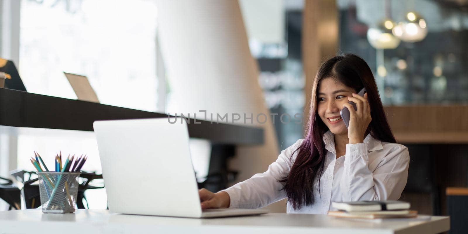 Young asian businesswoman beautiful charming smiling and talking on the mobile phone during typing on laptop computer in the office by nateemee