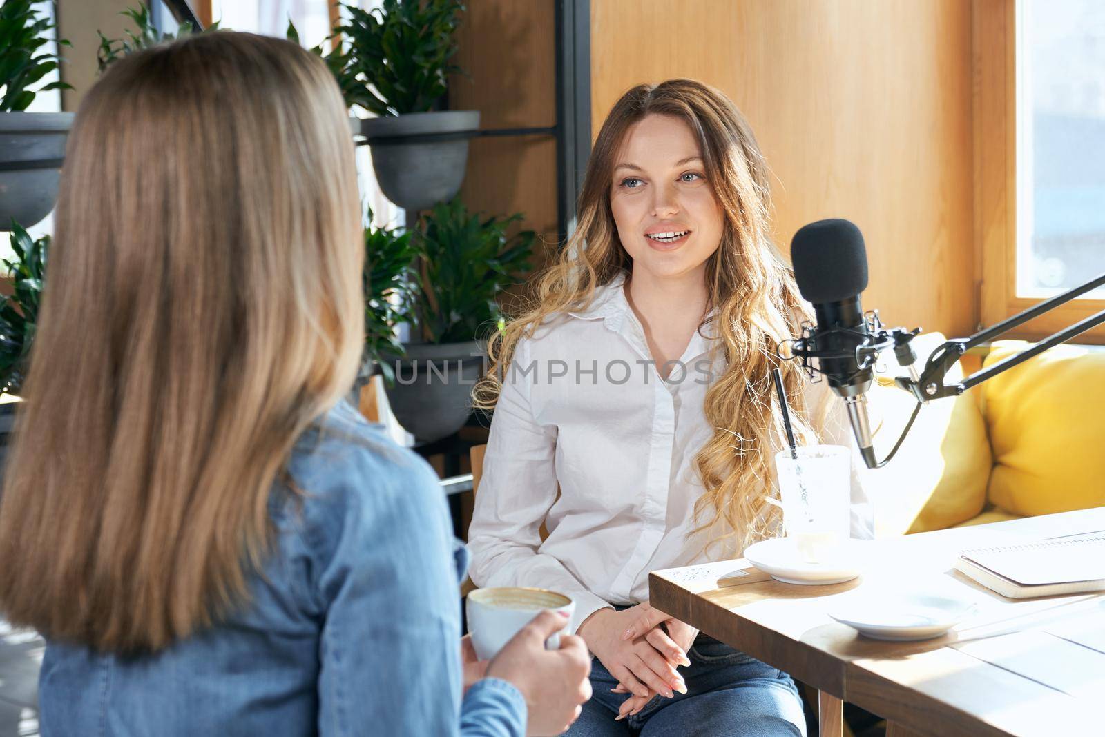 Close up young attractive women communicating different topics with cup coffee with modern black microphone. Concept of process interview with blogger woman in cafe with good mood.