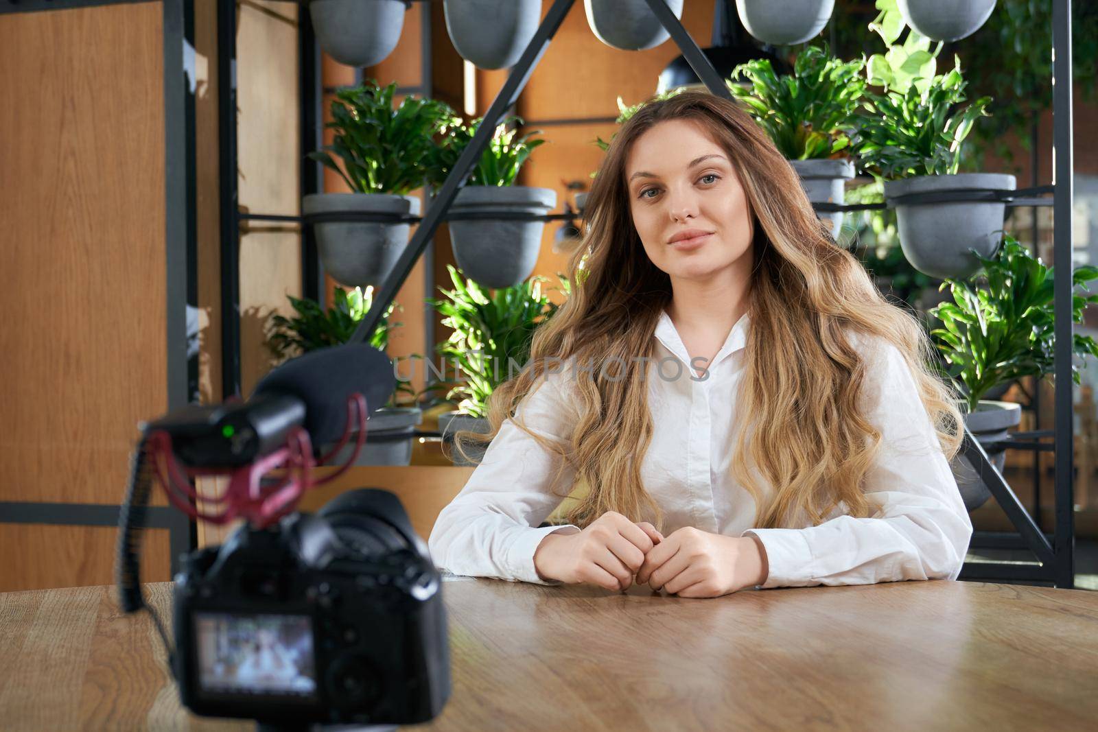 Front view of young beautiful woman blogger with long hair sitting at the table in cafe and preparing for interview on camera. Concept of process interview in modern cafe. 