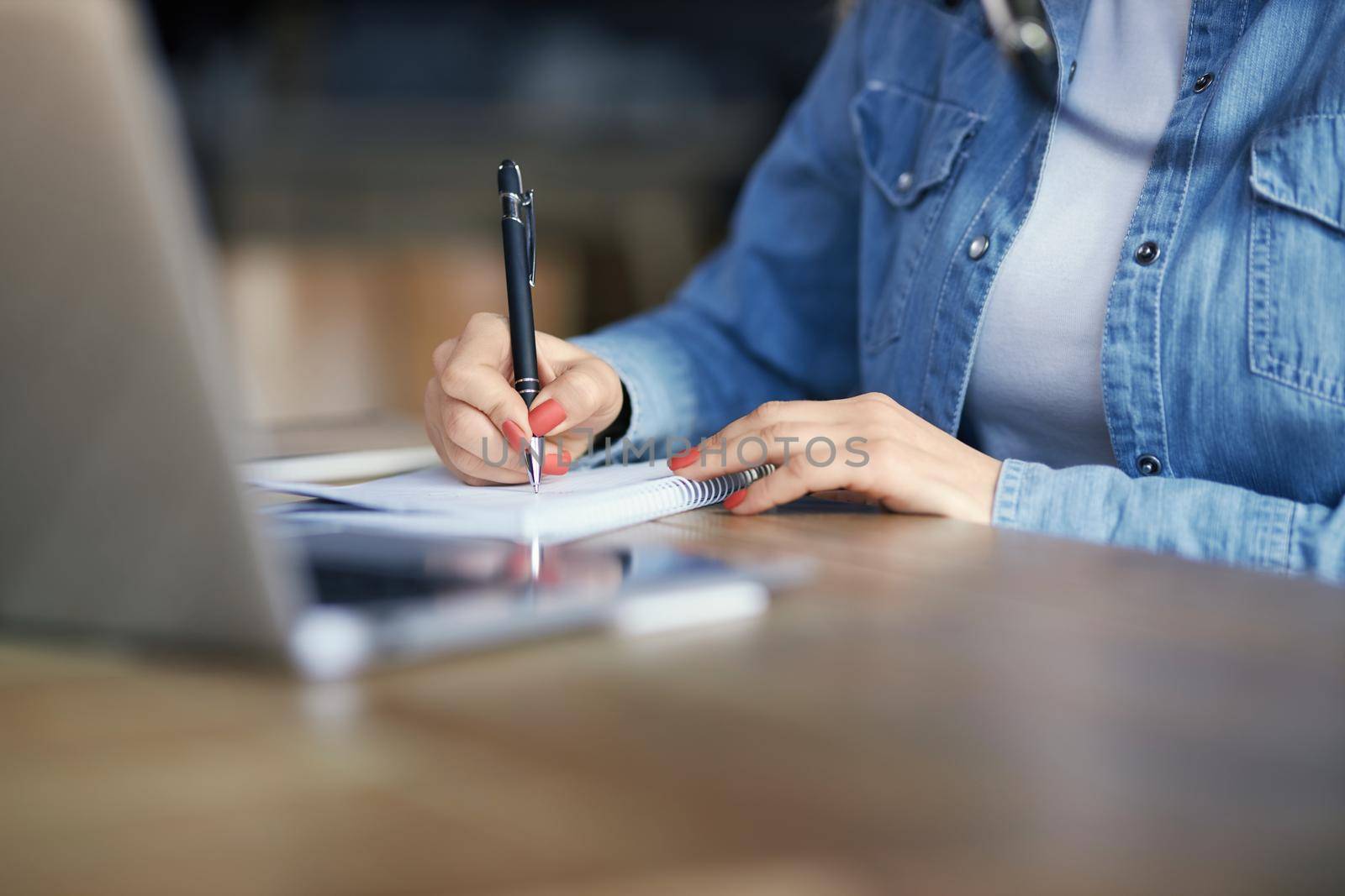 Close up of woman in denim jacket holding pen and writing important information in notebook. Concept of process preparing for communication about interesting facts. 