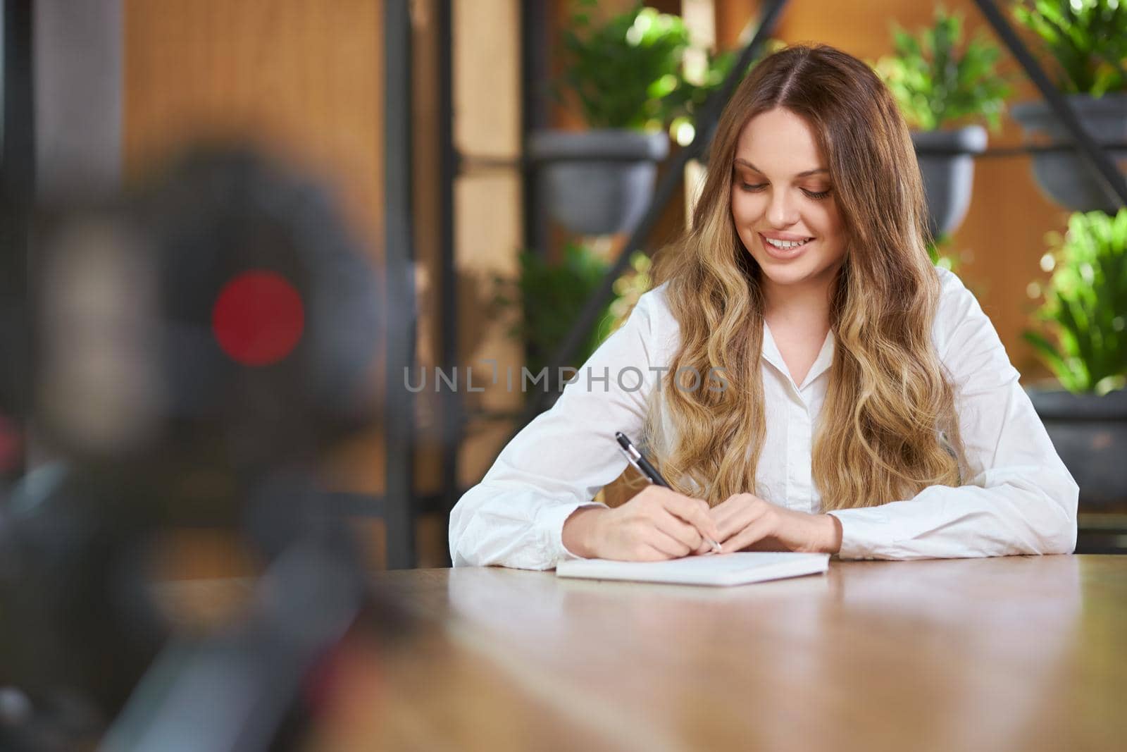 Front view portrait of smiling pretty young woman with long hair in white shirt sitting ar the table and working with notebook and pen. Concept of process working with good mood in cafe. 