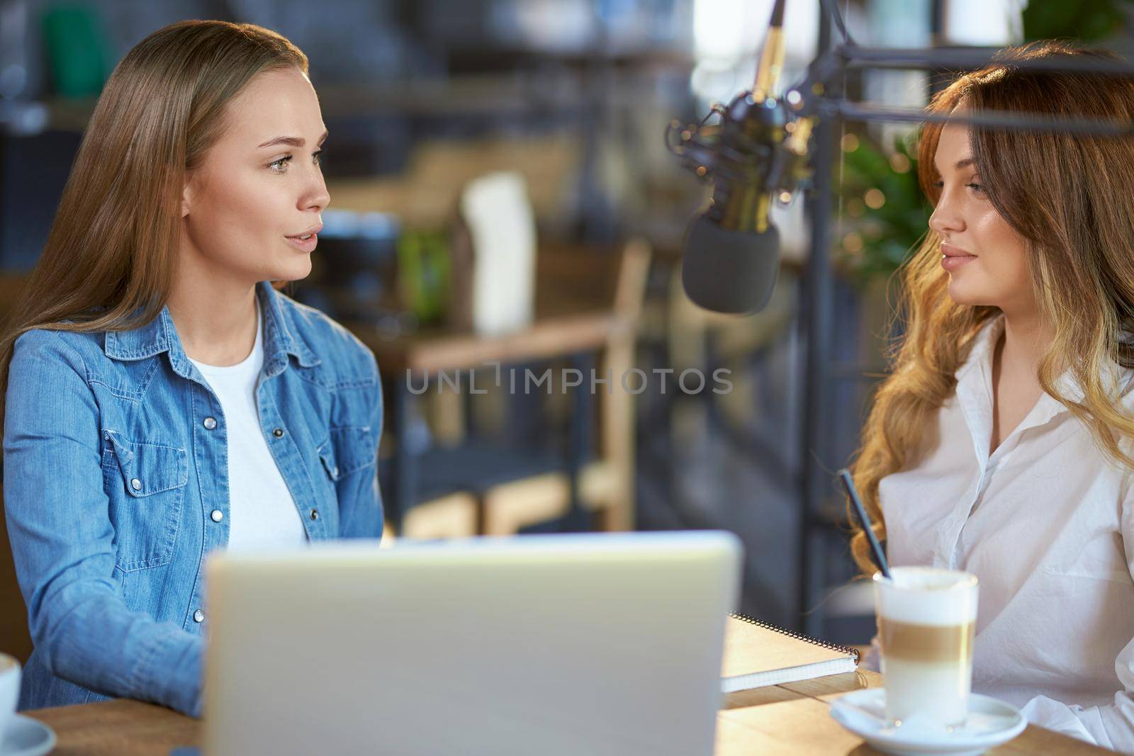Side view of two beautiful young blogger women communicating online with followers or friends in cafe with modern microphone. Concept of process interview with tasty coffee. 