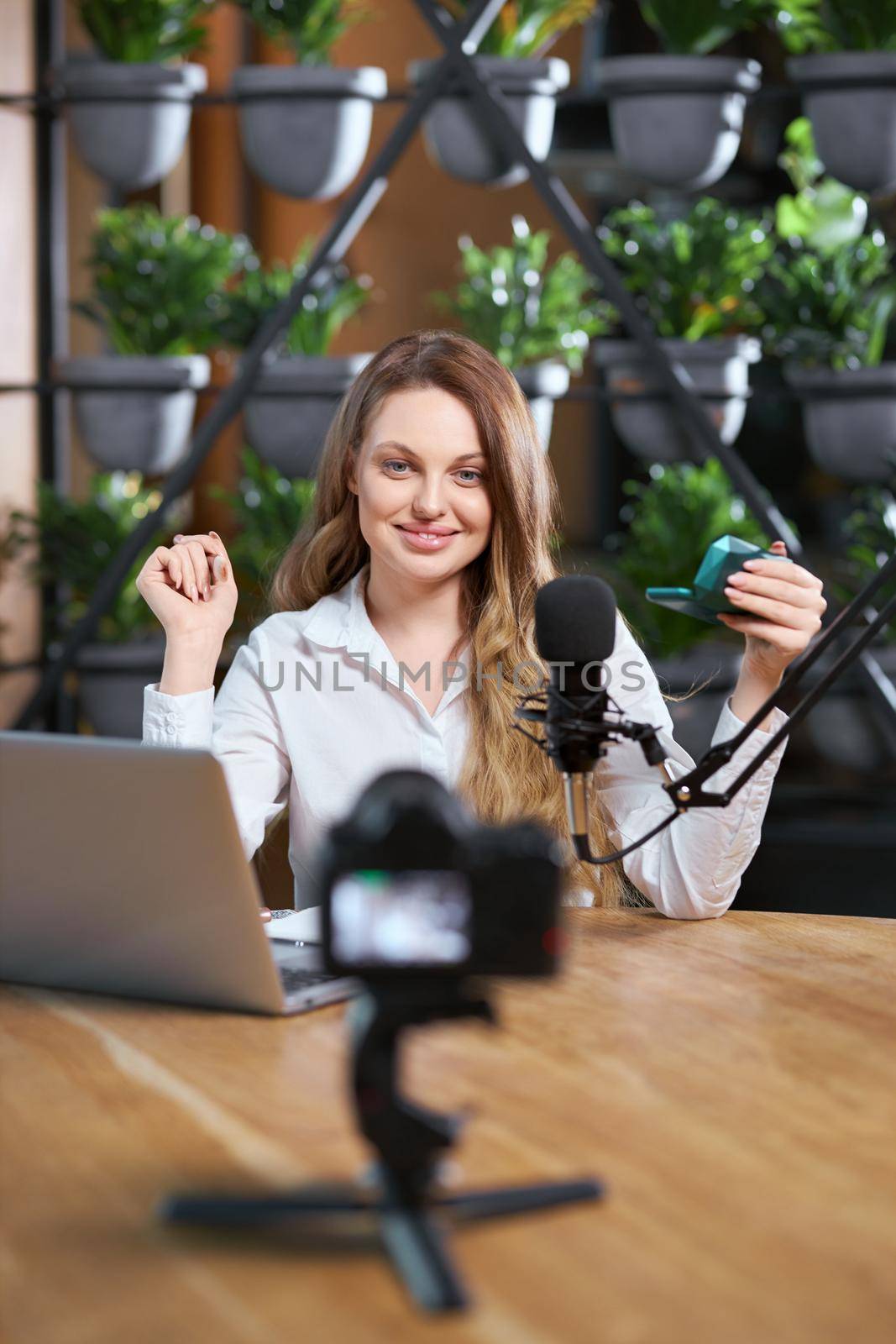 Front view of smiling young beautiful blogger woman in white shirt doing makeup at the table near special music equipment. Concept of process preparing for talking with followers in live broadcast. 