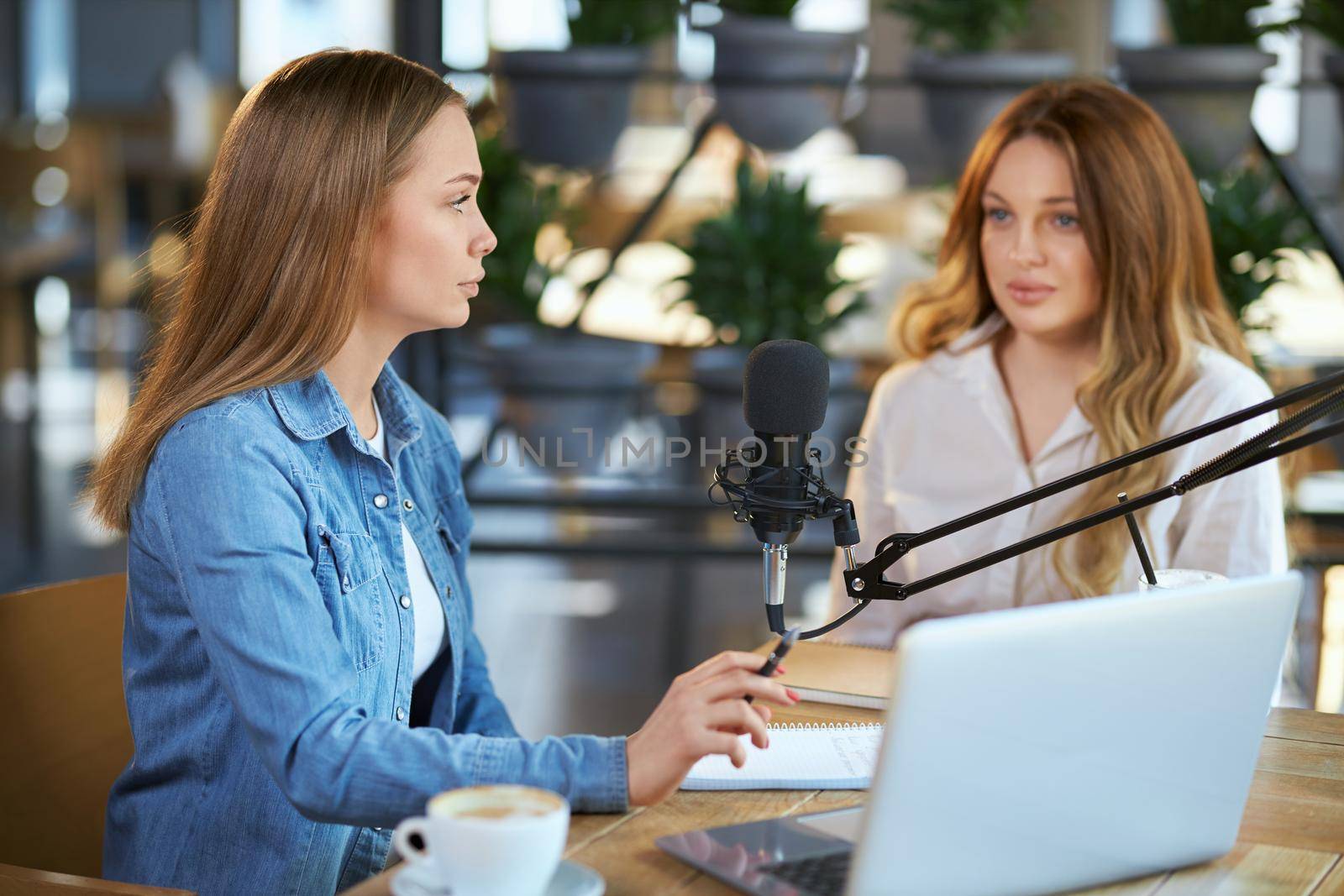 Side view of young attractive woman asks blogger question with modern microphone and laptop. Concept of process communication in cafe with tasty coffee and notebook,pen.
