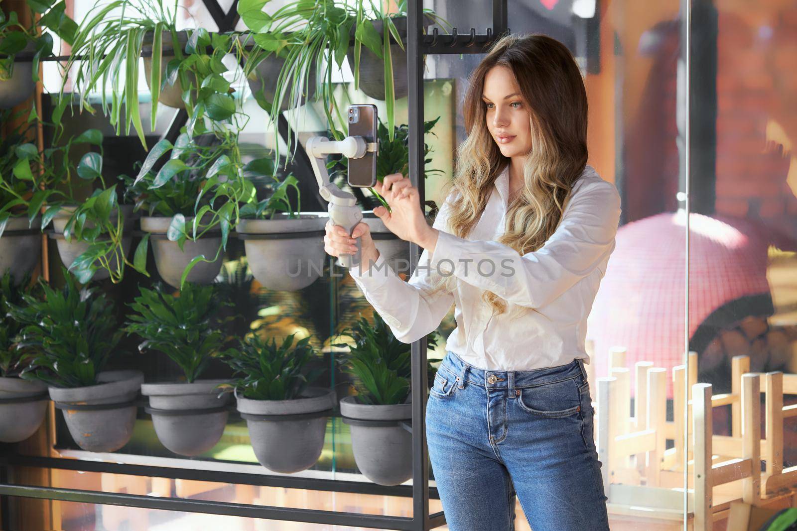 Side view of beautiful young woman in white shirt standing with selfie stick and doing selfie. Concept of process communicating with followers online on phone. 