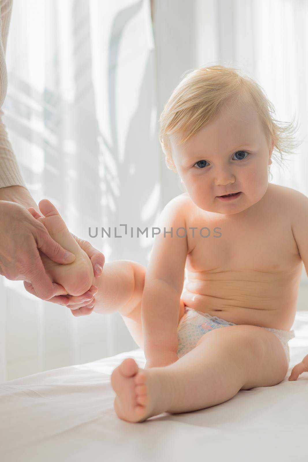 Mom gives her baby a leg and foot massage. Close-up. A satisfied baby is sitting on a massage table.