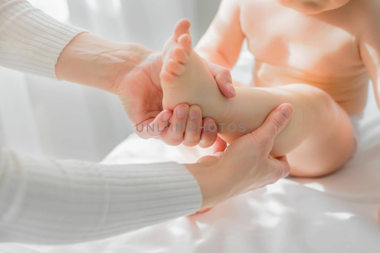 Mom gives her baby a leg and foot massage. Close-up. A satisfied baby is sitting on a massage table.