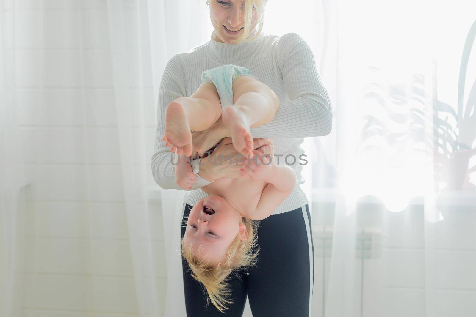 Mom holds the baby upside down, is engaged in children's gymnastics. Both are happy and laughing.