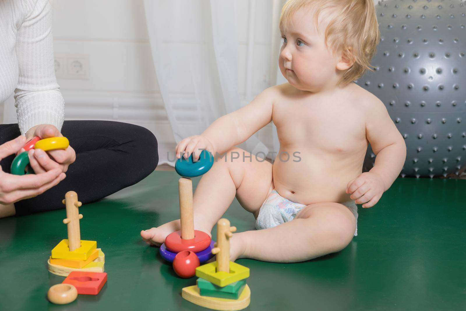 The kid sits on the rug, plays with his mother, collects a pyramid. Close-up.