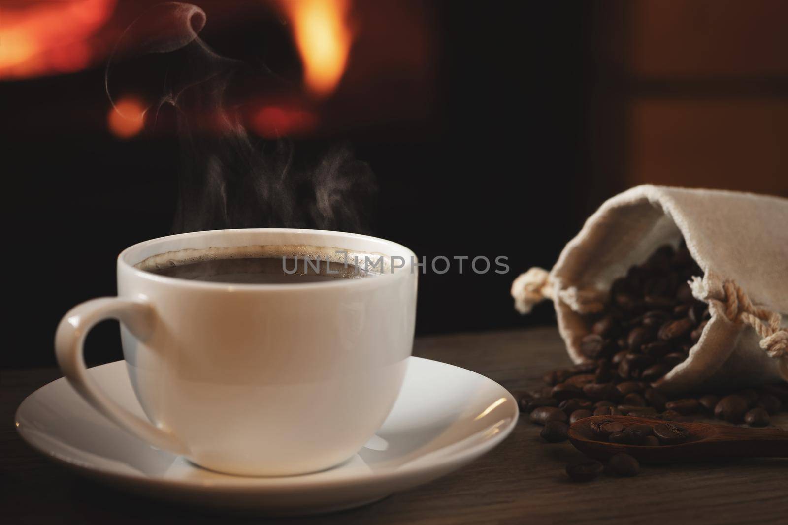 Close-up of cup of hot coffee and coffee beans in a bag on a wooden table in front of a burning fireplace. Selective focus.