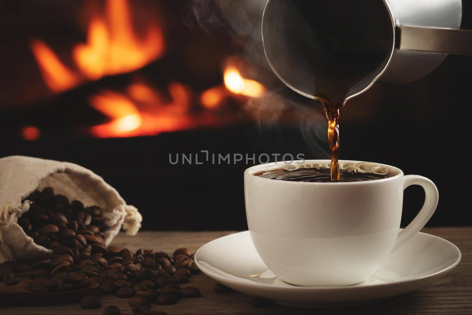 Pouring cezve coffee into a cup on a wooden table in front of a burning fireplace. Selective focus.