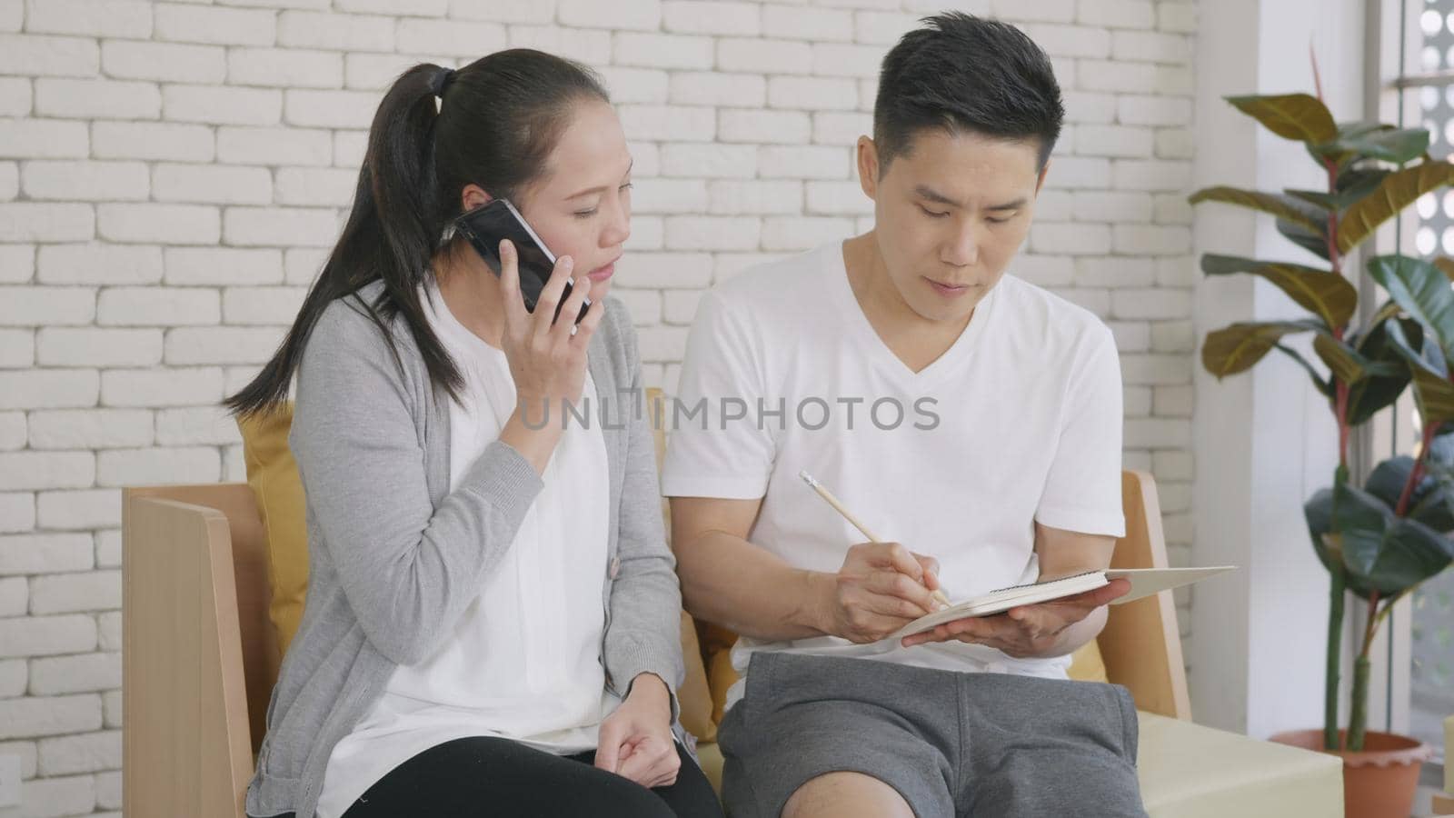 Happy Asian family couple husband and wife working confirming booking having talking call phone conversation and making writing notes of information together from home in living room.