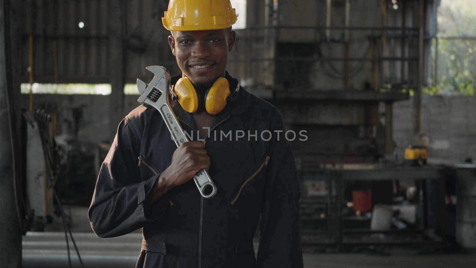 Portrait American industrial black young worker man smiling with helmet and ear protection in front machine, Engineer standing holding wrench on his shoulder at work in industry factory.