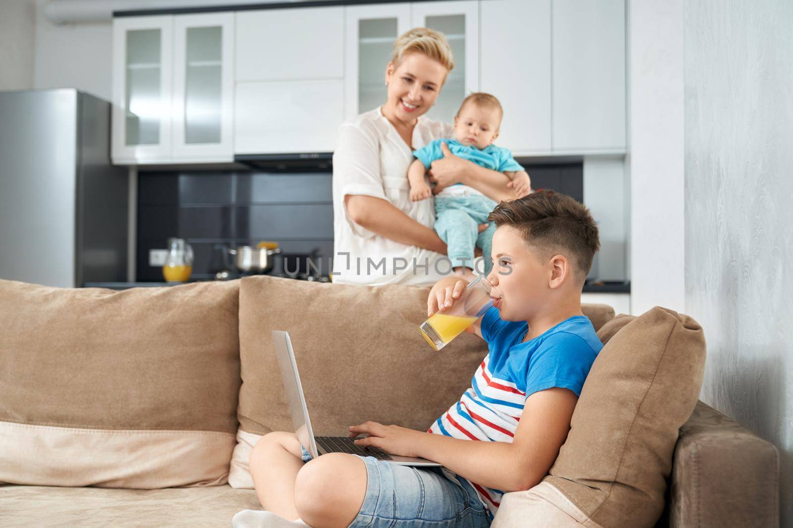 Happy boy drinking juice and sitting on couch with wireless laptop. Pretty mother playing with youngest son on kitchen. Family time.