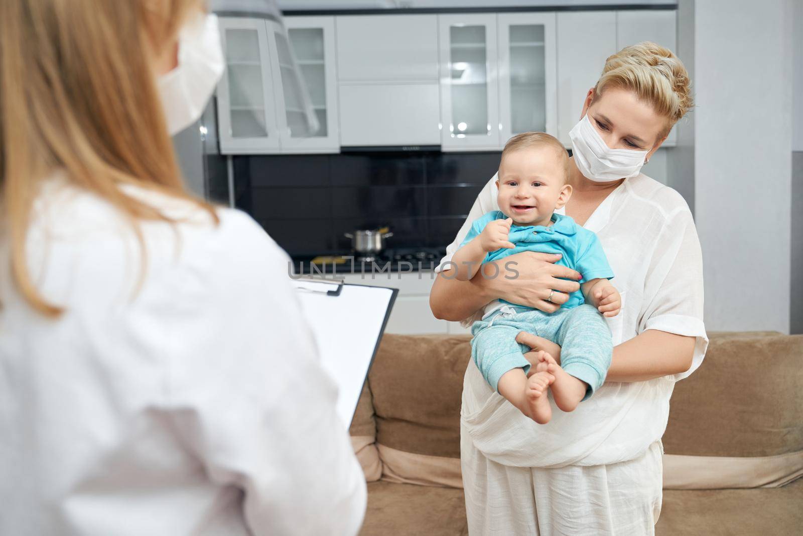 Female doctor writing something on clipboard and mother standing near and holding baby on hands. Pediatrician in protective mask visiting little patient at home.
