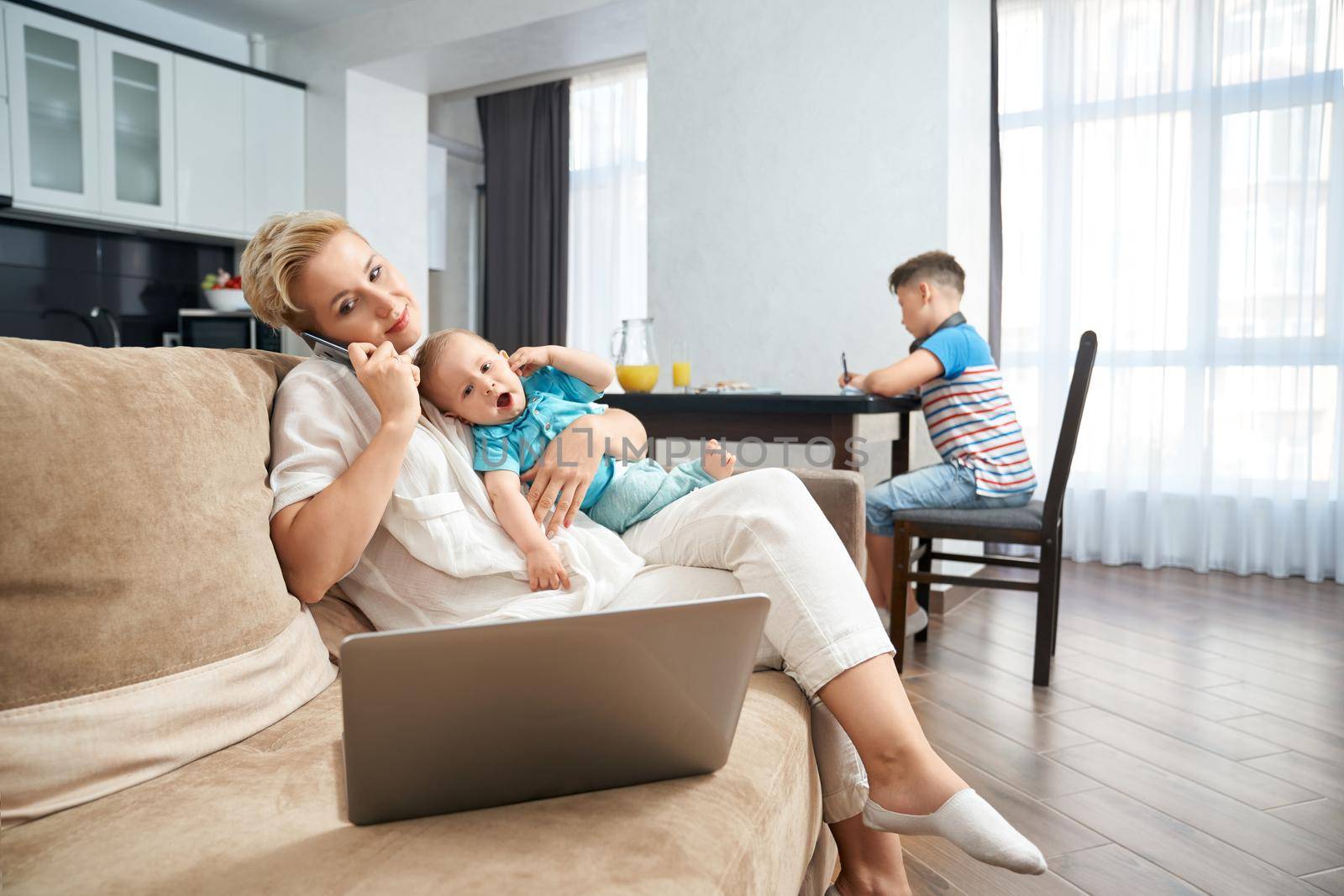 Beautiful woman talking on mobile, working on laptop and holding baby while her eldest son sitting at kitchen table and studying. Concept of motherhood and family time.