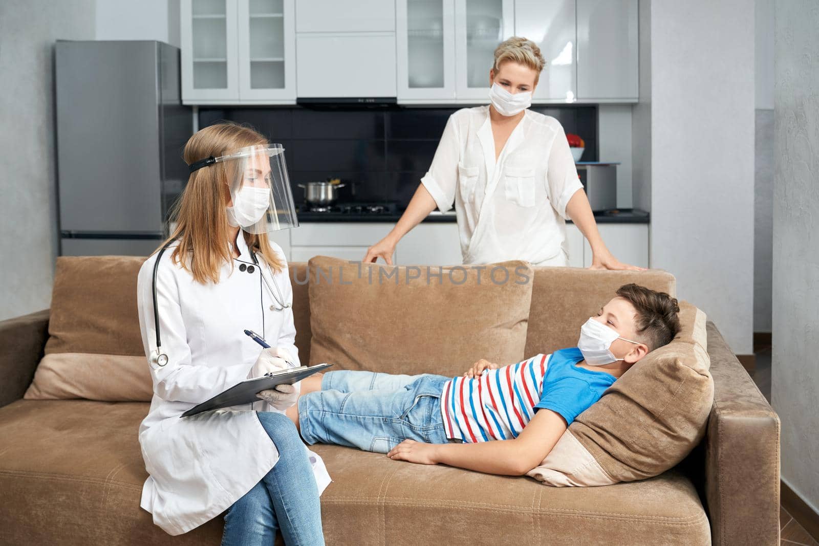 Competent pediatrician in protective mask and gloves writing on clipboard while doing checkup of teenager. Sick boy lying on couch and mother standing near at home.