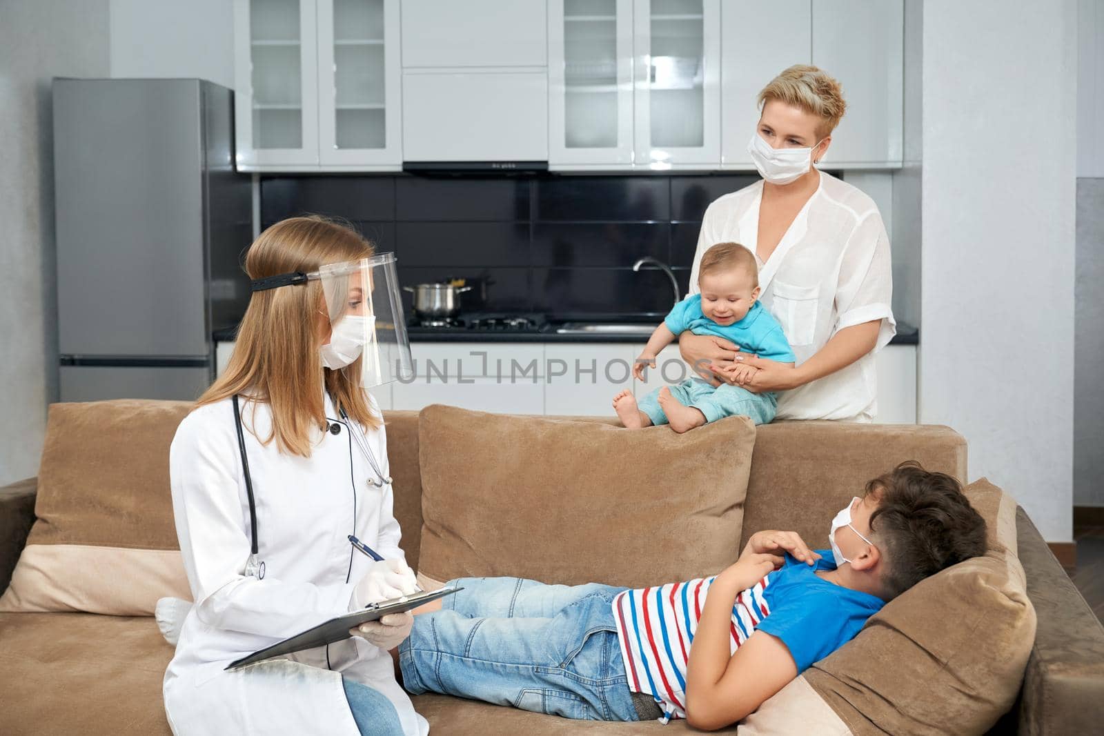 Female doctor in medical uniform examining sick teenager at home that lying on couch. Mother standing near with youngest son on hands. Pandemic time and children treatment.