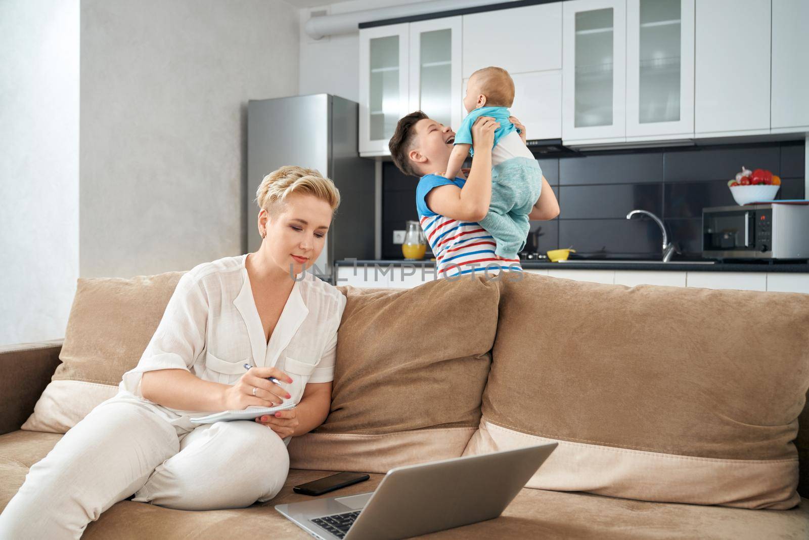Pleasant woman with short blond hair sitting on couch and working on laptop. To happy boys playing and smiling on kitchen. Concept of household and work.