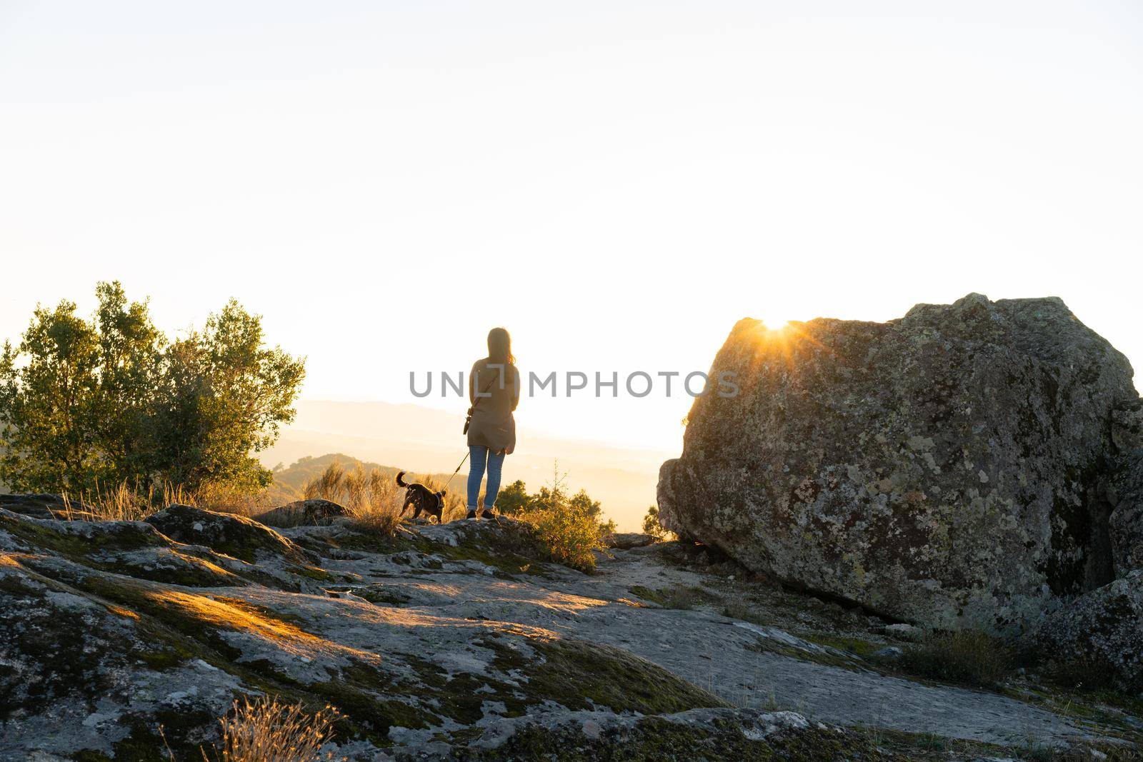 Caucasian young woman with brown dog seeing Sortelha nature mountain landscape with sun flare on a boulder, in Portugal by Luispinaphotography