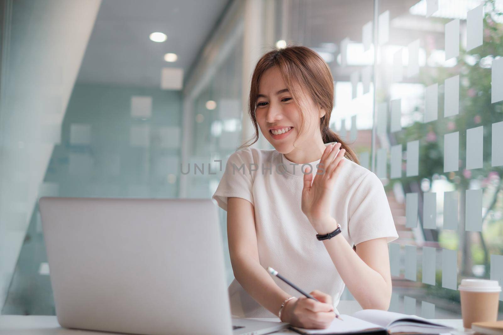 Happy Asian young business woman looking at computer screen, waving hello. using meeting online app, work from home, working remotely, social distancing, new normal concept. by itchaznong