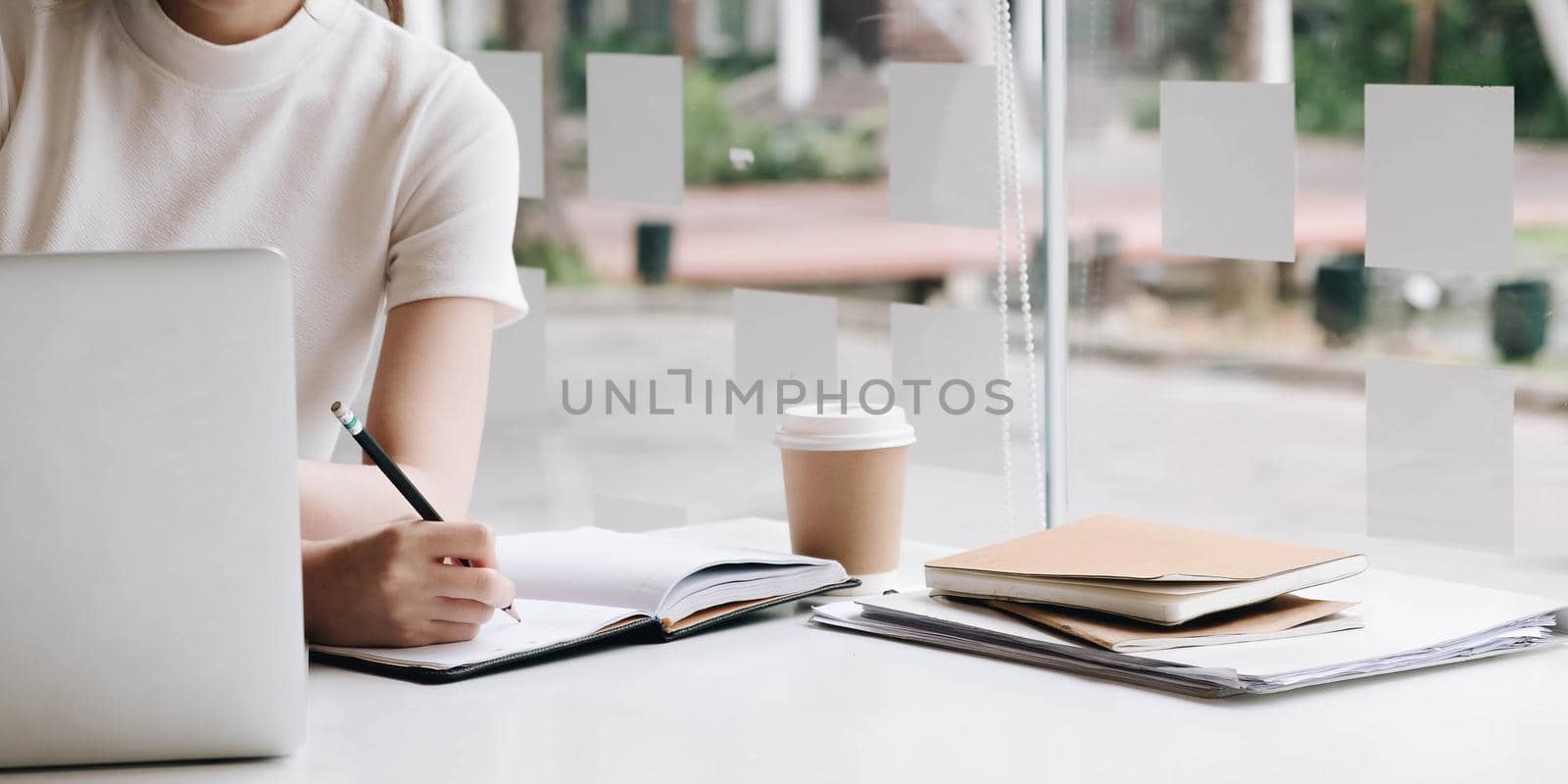 Business woman hand writing on notebook, work on laptop computer with calculator on the desk at modern home office. Online working, work at home concept by wichayada