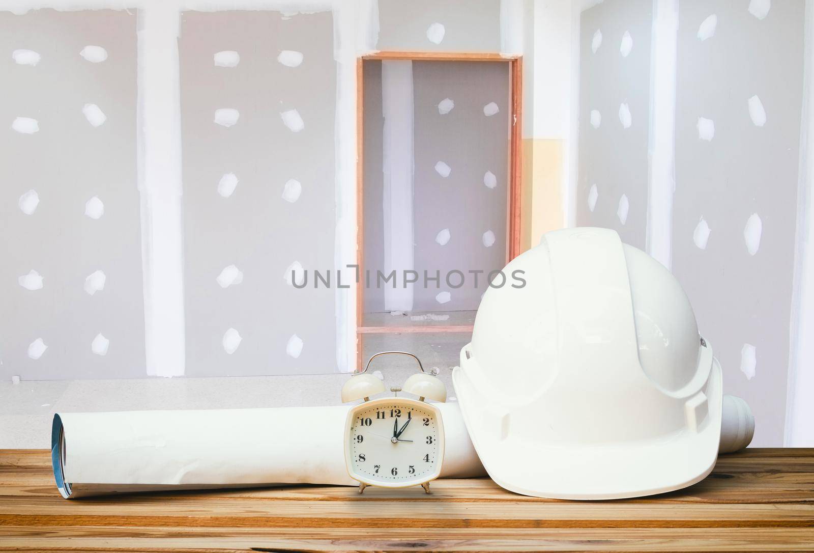 white safety helmet plastic, paper roll plan blueprint alarm clock time a rest at noon on wood floor table and gypsum board wall remodel interior with copy space add text