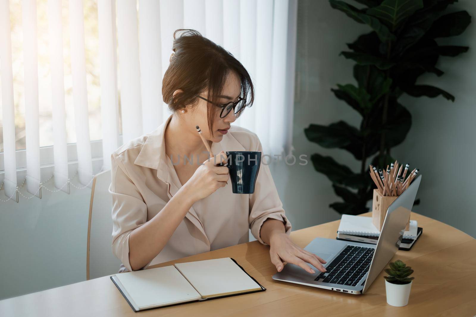 Portrait of beautiful accountant sitting at desk with interior drinking hot beverage holding cup with coffee looking at laptop while video conference. by itchaznong