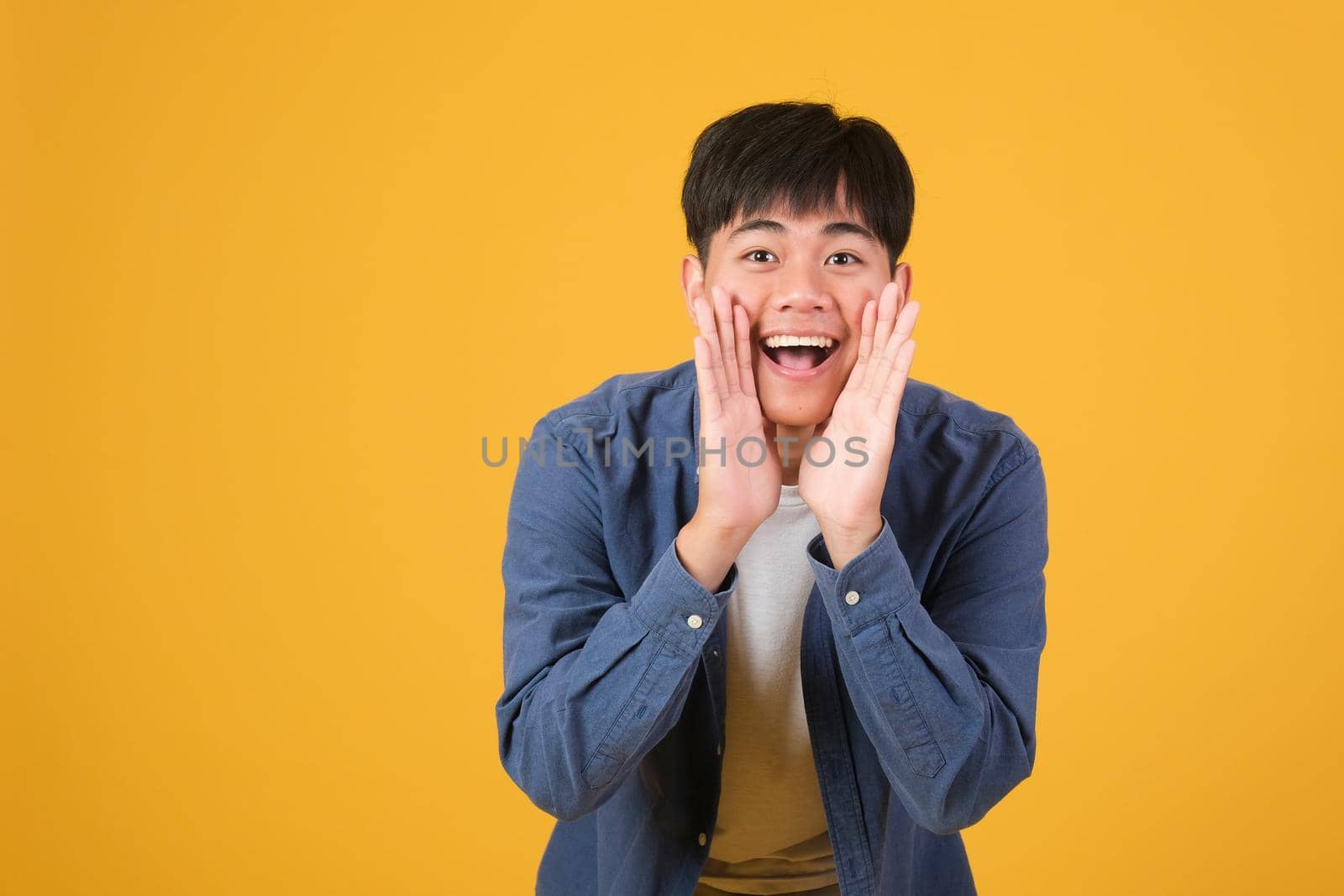 Portrait of excited asian young man hold hand mouth scream shout isolated on yellow orange color background.