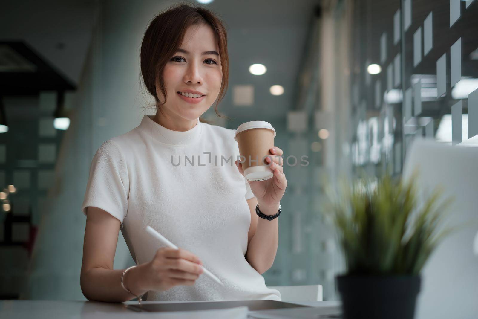 Cretive woman in white dress smiling holding stylus pencil and coffee in home office. Work from home concept