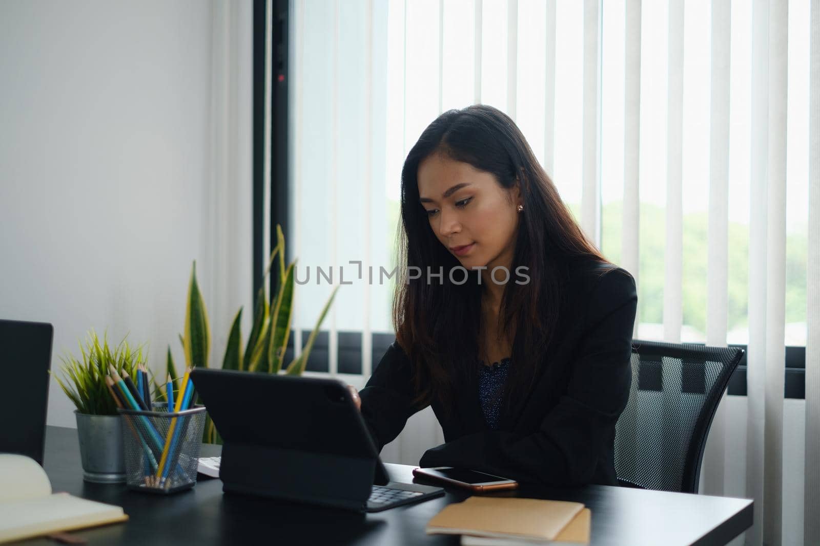 Portrait of beautiful accountant sitting at desk with interior drinking hot beverage holding cup with coffee looking at digital tablet while video conference