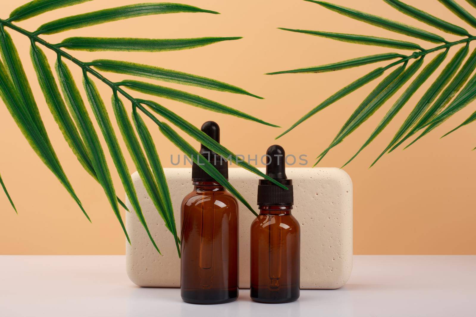 Two skin serum in dark brown bottles against beige podium and beige background with palm leaves. Concept of anti aging serum with natural ingredients 